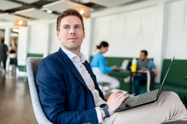 Collega met laptop op schoot en twee collega's op de achtergrond.